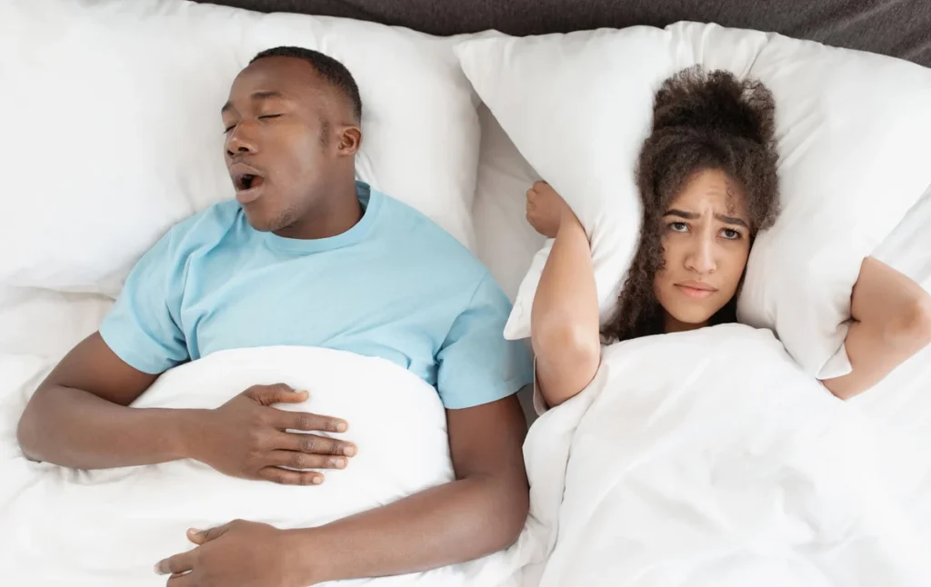 Snoring man sleeping with apnea and sleepless woman, angry wife unable to get to sleep. Upset young african american lady covers ears with pillow so as not to hear noise from snoring guy in bedroom
