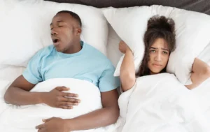 Snoring man sleeping with apnea and sleepless woman, angry wife unable to get to sleep. Upset young african american lady covers ears with pillow so as not to hear noise from snoring guy in bedroom